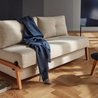 Sofá cama Cubed Wood bege com pernas de madeira e manta azul.