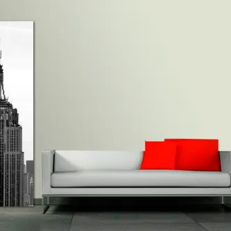 Quadro vertical Empire State em preto e branco junto a sofá branco com almofadas vermelhas