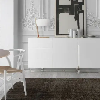 Aparador Ellena branco minimalista com três gavetas, candeeiro e quadro numa sala.