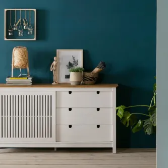 Aparador Bruna branco com gavetas e porta ripada, decoração sobre o tampo e parede azul-petróleo