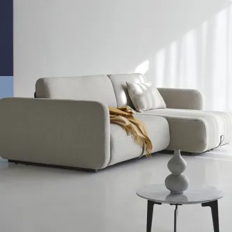 Sofá cama Vogan cinzento claro com chaise, almofada e manta numa sala minimalista