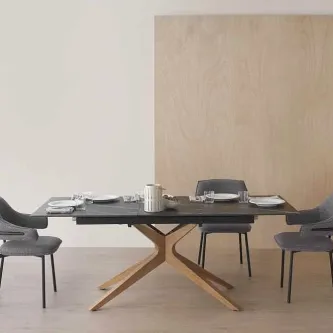 Mesa extensível com tampo cerâmico escuro e base em madeira em X, seis cadeiras cinzentas e loiça.