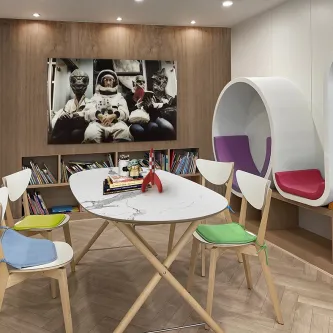 Quadro galaticos com astronauta e figuras extraterrestres numa sala com mesa e cadeiras coloridas