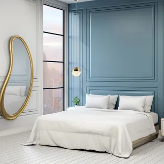 Espelho Brigitte oval dourado encostado na parede num quarto com cama branca