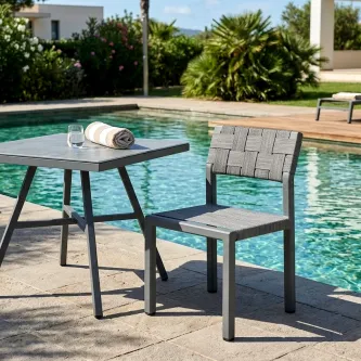Mesa de jardim quadrada cinzenta junto à piscina com cadeira entrelaçada e toalha