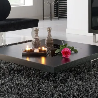 Mesa de centro preta baixa com velas, castiçais prateados e rosa sobre tapete cinzento.