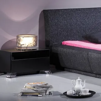 Mesa de cabeceira silver preta com pernas prateadas e candeeiro iluminado ao lado de cama