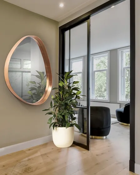 Espelho Julie redondo com moldura cobre na parede, planta em vaso no chão