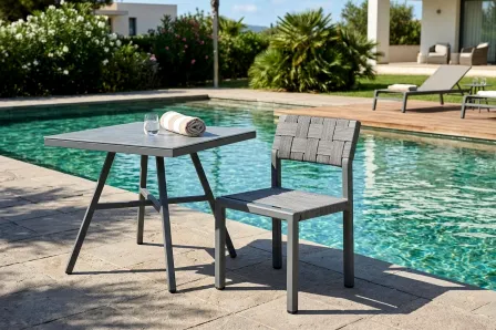 Mesa de jardim quadrada cinzenta junto à piscina com cadeira entrelaçada e toalha
